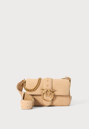 Beige suède handtas met een voorflap, goudkleurige hardware, kettingriem en een decoratieve ring met vogelaccenten. Bevat studs op de flap.