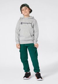 Ragazzo che indossa un hoodie grigio della Champion, pantaloni cargo verdi, sneakers nere e bianche e un cappellino verde indossato all'incontrario, in piedi davanti a uno sfondo bianco.