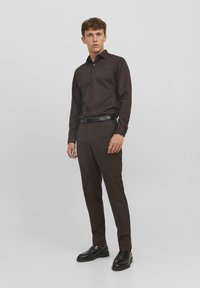 Camicia button-up marrone scuro con maniche lunghe, abbinata a pantaloni a quadretti marroni e mocassini neri. Vestibilità strutturata, tessuto morbido, design minimalista.
