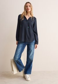 Marineblauwe blouse met lange mouwen en een V-hals, gecombineerd met lichtblauwe wijde jeans en witte sneakers, en een crèmekleurige tas in de hand.