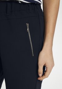 Mano destra appoggiata accanto a pantaloni neri con una tasca zipper verticale argentata, indossati con una camicia a righe.