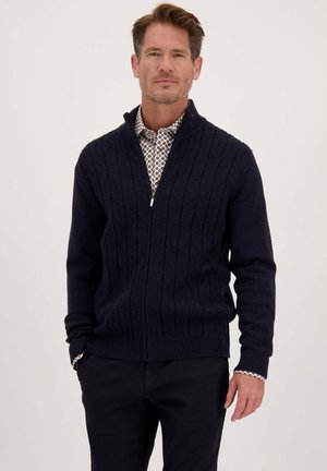 Man in een marineblauwe kabelgebreide ritssweater over een gestippeld shirt, staande met de handen in de zakken tegen een effen achtergrond.