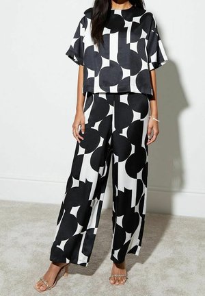 Kvinde iført en sort og hvid løstsiddende top med geometrisk mønster og vidde bukser med åbne hæle, stående op ad en hvid væg.