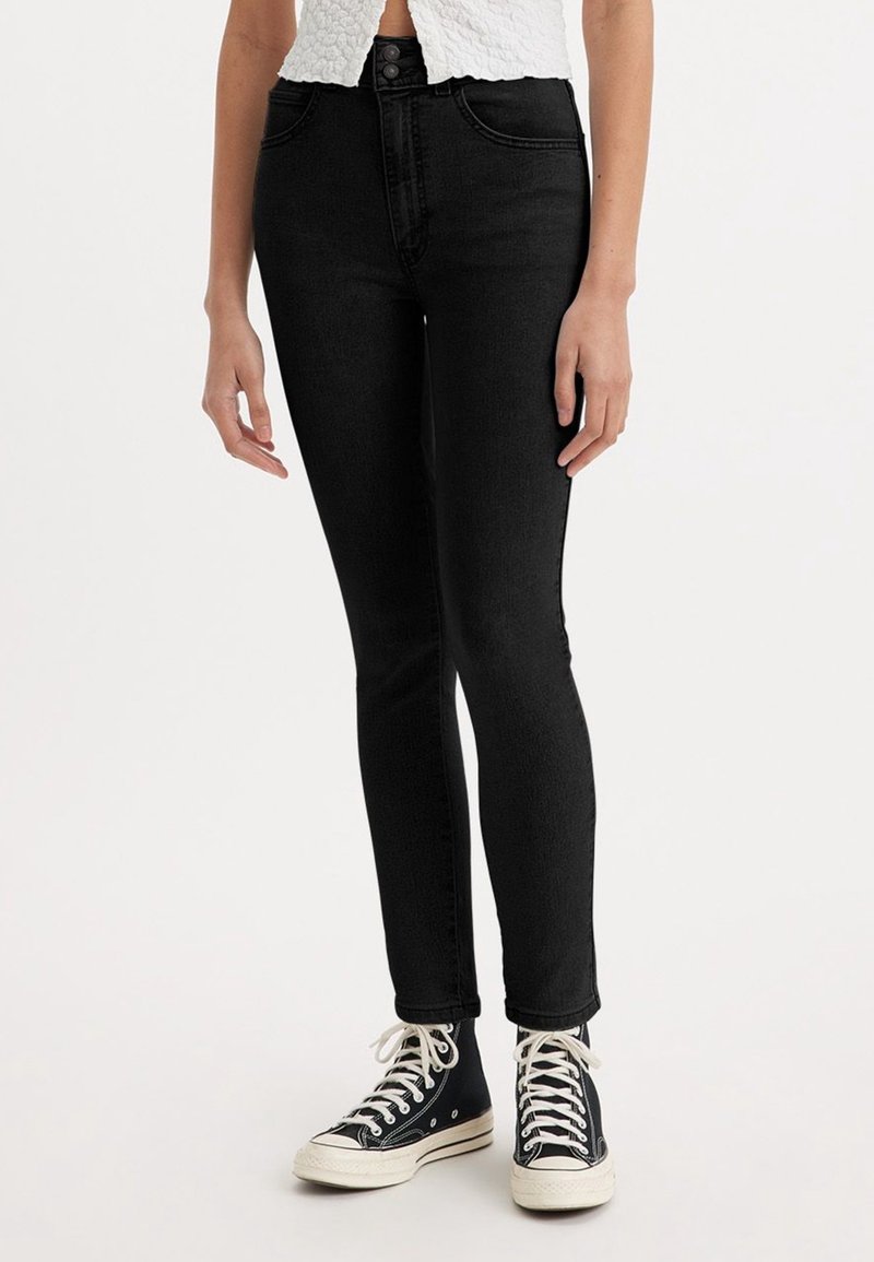Jean skinny noir taille haute, en denim. Comprend des passants de ceinture et un design classique à cinq poches. Porté avec des baskets montantes noires.