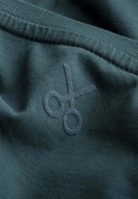 Donkergroene stof met een gestructureerde afwerking heeft een fletse, geborduurde schaarlogo in een contrasterende kleur. Schoon ontwerp, geen andere details zichtbaar.