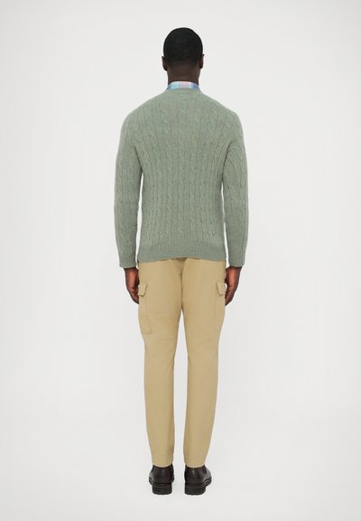 Camisola verde de malha com padrão de cabo, calças cargo bege e sapatos escuros, vistos de trás. A camisola apresenta punhos e barra canelados.