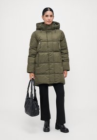 Mujer vestida con un abrigo acolchado verde oliva con capucha, pantalones negros, botas de plataforma y sosteniendo un bolso negro sobre un fondo blanco liso.