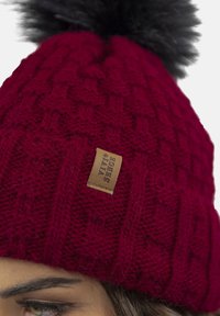Berretto rosso lavorato a maglia con un motivo testurizzato e un pompon in pelliccia nera sulla sommità. Presenta un piccolo etichetta in pelle marrone sul lato.