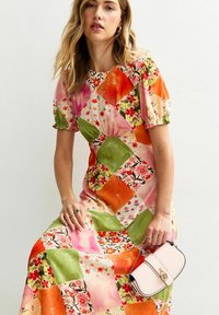 Kvinna som bär en färgglad lapptäcks-klänning med blommiga och enfärgade rutor i orange, rosa och grönt, hållande en krämfärgad handväska med ett gyllene lås.