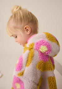 Veste à capuche en polaire avec motifs abstraits roses, jaunes et verts. Texture douce et bords arrondis. La capuche repose doucement sur le dos.