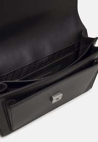 Svart läderhandväska med en slät yta, utrustad med en zipficka, en logotyp och en magnetisk stängning. Inuti har den ett vadderat mönster.