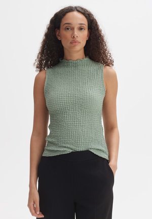 Hellgrünes ärmelloses Oberteil mit einem strukturierten, gekräuselten Design und einem gerafften hohen Kragen. Kombiniert mit schwarzen, eng anliegendem Hosen.