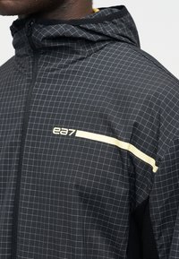 Άνδρας που φοράει μαύρο μπουφάν με κουκούλα με λευκό σχέδιο πλέγματος, φερμουάρ μπροστά και λογότυπο "ea7" με οριζόντια ρίγα στο στήθος και το μανίκι.