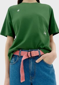 Groene katoenen t-shirt met korte mouwen, versierd met een geborduurd witte bloem en de tekst "sot". Gecombineerd met hoge blauwe spijkerbroeken en een roze gevlochten riem.