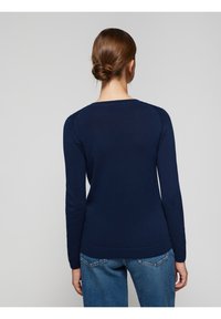 Námořnicky modrý svetr s dlouhým rukávem, kulatým výstřihem a žebrovaným lemem, s hladkou texturou a přiléhavým střihem. Kombinován s světlými džínami z denimu.