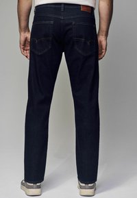 Vaqueros de mezclilla azul oscuro con corte recto, que cuentan con dos bolsillos traseros, costuras prominentes y un parche de cuero en la cintura.