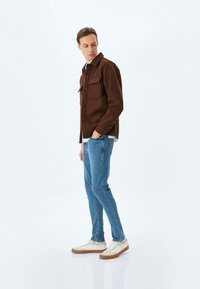 Chaqueta marrón de botones con dos bolsillos en el pecho, combinada con jeans ajustados azules y zapatillas blancas con detalles marrones, de pie sobre un fondo simple.