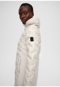 Pufferjacke mit einer cremefarbenen gesteppte Außenschicht, ausgestattet mit einer hellbeigen Kapuze und Seitenpanelen. Schwarzes Logo-Patch am linken Arm.