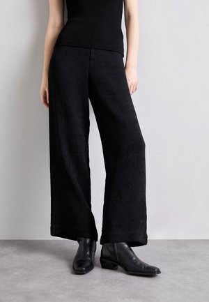 Femme portant un pantalon noir à jambes larges, un haut sans manches noir et des bottines noires, debout sur un sol gris contre un mur gris clair.