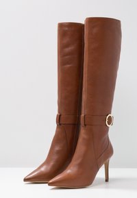 Bottes hautes jusqu'au genou en cuir marron avec bouts pointus, fermeture éclair à l'avant et accent boucle à la cheville. Talons aiguilles pour une touche d'élégance.