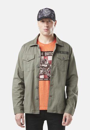 Homme portant une casquette noire avec un logo blanc, une veste boutonnée vert olive sur un T-shirt graphique orange avec un motif de moto, debout devant un fond blanc.