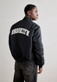 Crna bomber jakna s bijelim vezom "BROOKLYN" na leđima, s rebrastim ovratnikom i uskim manžetama. Uparena s tamnim trapericama.