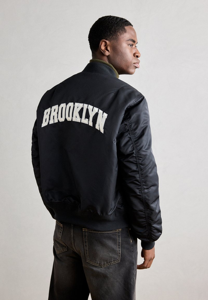 Crna bomber jakna s bijelim vezom "BROOKLYN" na leđima, s rebrastim ovratnikom i uskim manžetama. Uparena s tamnim trapericama.