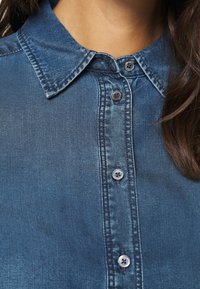 Blå denimskjorta med knapp-down krage, med ljusblå knappar och synliga sömnadsdetaljer längs knappslån och kragens kanter.