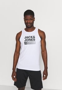 Débardeur blanc avec un logo noir en gras "JACK & JONES PERFORMANCE", offrant une coupe ajustée et un col rond, associé à un short noir.