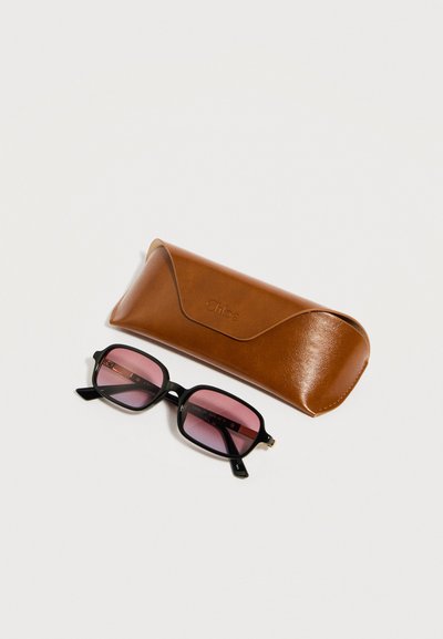 Μαύρα ορθογώνια γυαλιά ηλίου με ροζ φακούς δίπλα σε καφέ δερμάτινη θήκη της μάρκας Chloe σε λευκό φόντο.