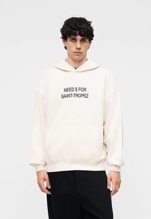 Νεαρός άνδρας με σγουρά μαλλιά που φοράει μια εκρού φούτερ με το κείμενο «ΧΡΕΙΑΖΟΜΑΙ ΧΡΗΜΑ ΓΙΑ ΣΕΝ-ΤΡΟΠΕ» σε συνδυασμό με μαύρο παντελόνι, μπροστά σε απλό φόντο.