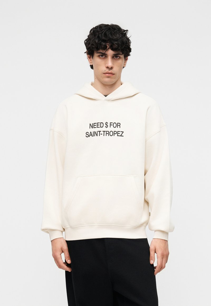 Jeune homme aux cheveux bouclés portant un sweat à capuche écru avec l'inscription « NEED $ FOR SAINT-TROPEZ », associé à un pantalon noir, devant un fond uni.