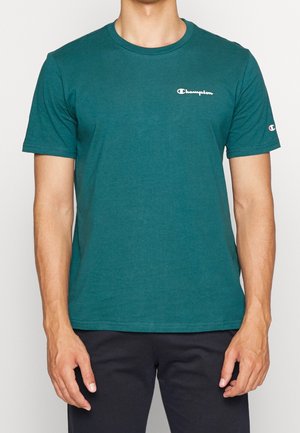 Mężczyzna w turkusowej koszulce Champion z logo na piersi i rękawie, w czarnych spodniach, stojący na jednolitym tle.