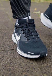 Shoe atlético preto com parte superior em malha, Swoosh branco da Nike, detalhes cinzentos e amortecimento Air Zoom visível numa sola texturizada.