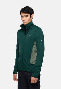 Giacca verde con zip e colletto alto, tessuto texturizzato e tasca laterale beige a contrasto. Presenta il logo sul petto e sulla spalla.