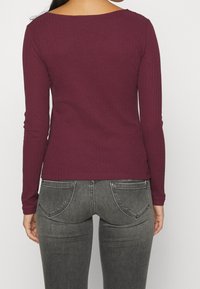 Burgunderfarbenes Langarmoberteil mit geripptem Muster und weitem Ausschnitt, kombiniert mit grauen Jeans mit Gesäßtaschen und einem faded Look.