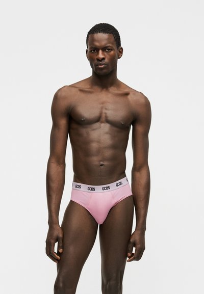 Μυώδης μαύρος αρσενικός μοντέλο που φοράει ροζ εσώρουχα GCDS, στέκεται μπροστά σε απλό ανοιχτό γκρι φόντο, με ουδέτερη έκφραση και βλέμμα προς τα εμπρός.