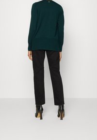 Persona con un suéter verde oscuro, pantalones rectos negros y botines de tacón alto negros, de pie contra un fondo blanco liso.
