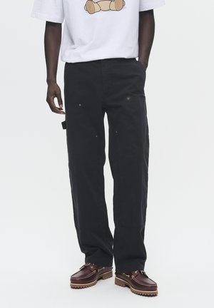 Pantalones cargo negros con múltiples bolsillos, costuras visibles y un diseño de pierna recta. Llevados con una camiseta gráfica blanca y zapatos marrones.