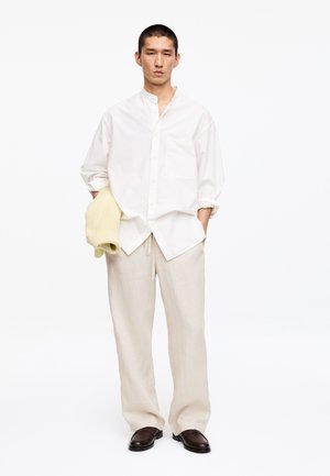 Homme portant une chemise blanche ample à boutons, un pantalon beige à jambes larges, des chaussures marron foncé, tenant un pull crème plié dans une main.