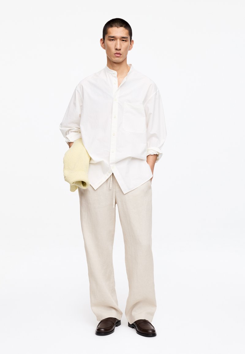 Homme portant une chemise blanche ample à boutons, un pantalon beige à jambes larges, des chaussures marron foncé, tenant un pull crème plié dans une main.
