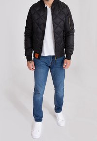 Veste bomber noire matelassée avec poignets et ourlet côtelés, poche zipp ée sur la manche, portée avec un jean bleu et des baskets blanches.