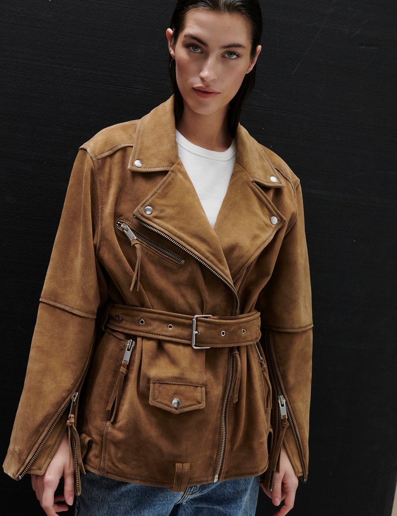 Γυναίκα με καφέ σουέντ μπουφάν με ζώνη, φερμουάρ στις τσέπες και ασημένια κουμπιά, φορεμένο πάνω από λευκό πουκάμισο, στέκεται μπροστά σε μαύρο τοίχο.