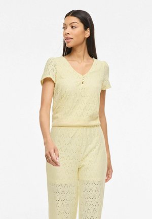 Femme aux longs cheveux foncés portant un haut en tricot à manches courtes jaune pâle et un pantalon assorti avec un motif ajouré en forme de losange.