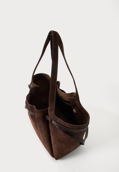 Sac cabas en daim marron avec deux fines bretelles d'épaule et un dessus ouvert montrant une poche intérieure zippée sur un fond clair uni.