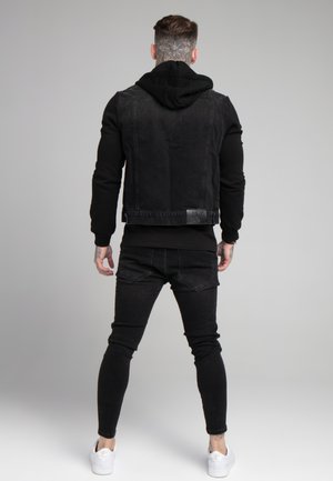 Mężczyzna z tatuażami na szyi i rękach, ubrany w całkowicie czarny strój z kapturem i białe trampki, zwrócony tyłem na szarym tle.