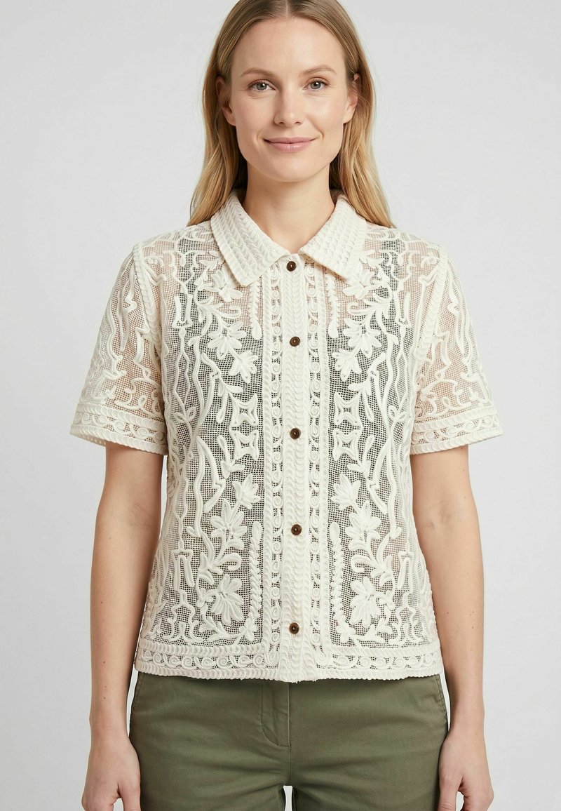 Femme portant un chemisier en dentelle à manches courtes de couleur crème avec des motifs floraux et des boutons marron, associé à un pantalon vert olive.