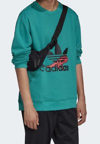 Teal sweatshirt dengan logo Adidas hitam menampilkan leher bulat dan manset bergaris. Tas selempang hitam yang dapat disesuaikan dikenakan di bahu.
