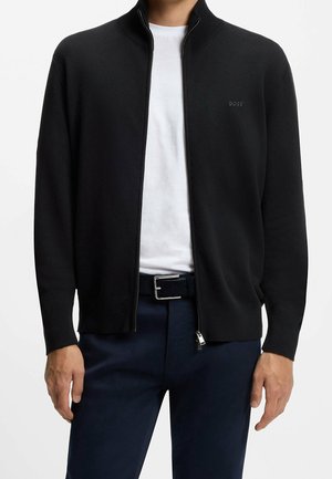 Homme portant une veste noire à fermeture éclair sur un t-shirt blanc et un pantalon bleu foncé avec une ceinture noire, debout devant un fond uni.