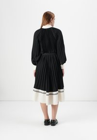 Model wearing a black pleated dress with a white collar and hem, standing against a plain white background, showcasing the garment's design.